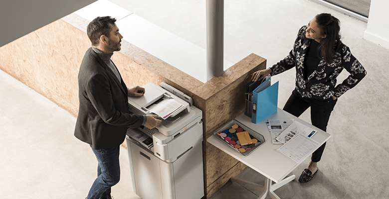 A man stood using a printer while talking to a female colleague in an office environment