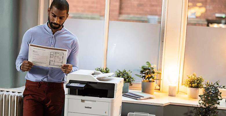 A man stood next to a printer, reading a document in an office environment