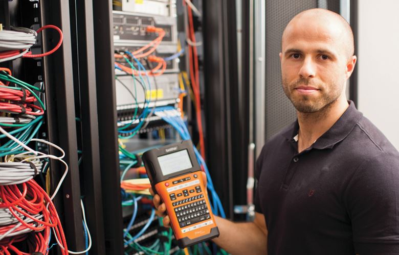 Male network technician stands in front of a neatly labelled network cabinet, whilst holding a P-touch label printer