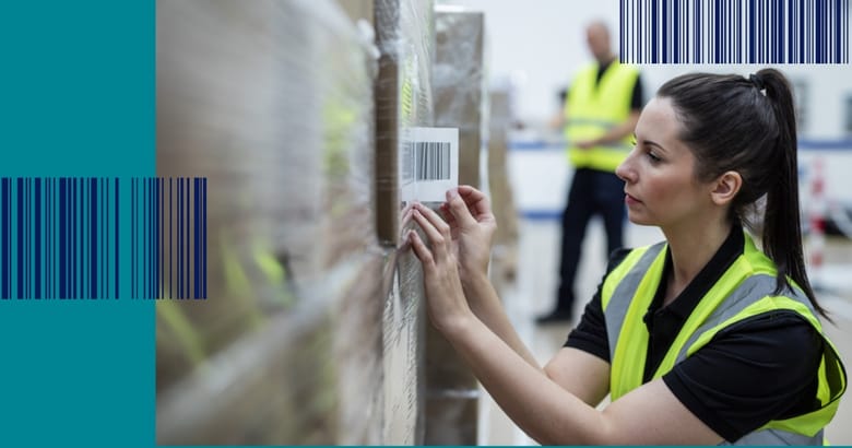 A female warehouse worker wearing a high visibility vest sticking a barcode label to a shrink-wrapped pallet of brown cardboard boxes
