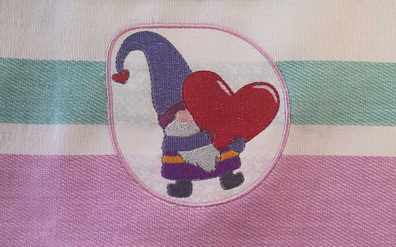 Embroidered gnome holding red heart on pink and green striped background 