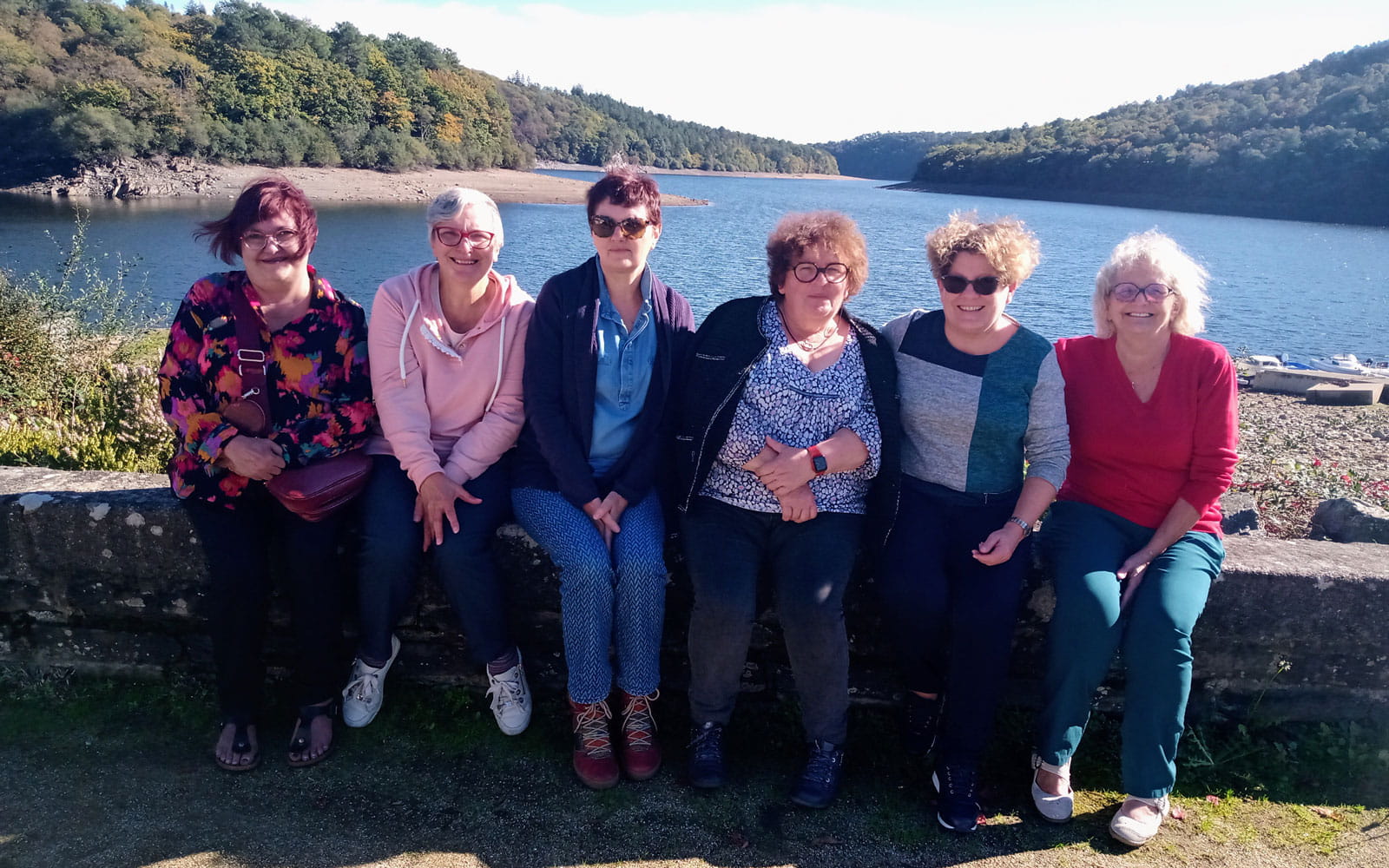 group of 6 women sitting on low wall in front of lake