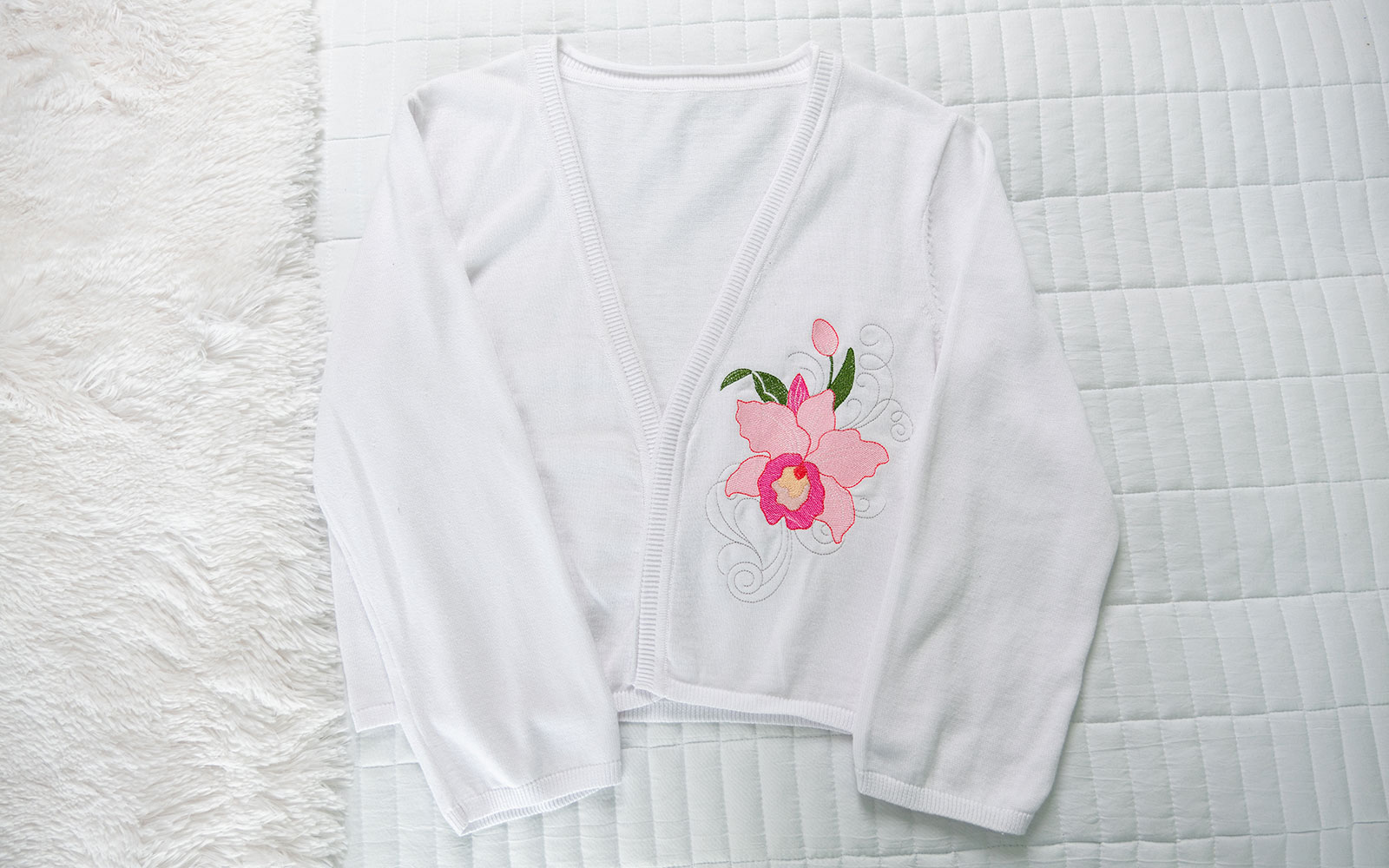 cardigan bianco con ricamo orchidea rosa sul petto disteso su letto bianco