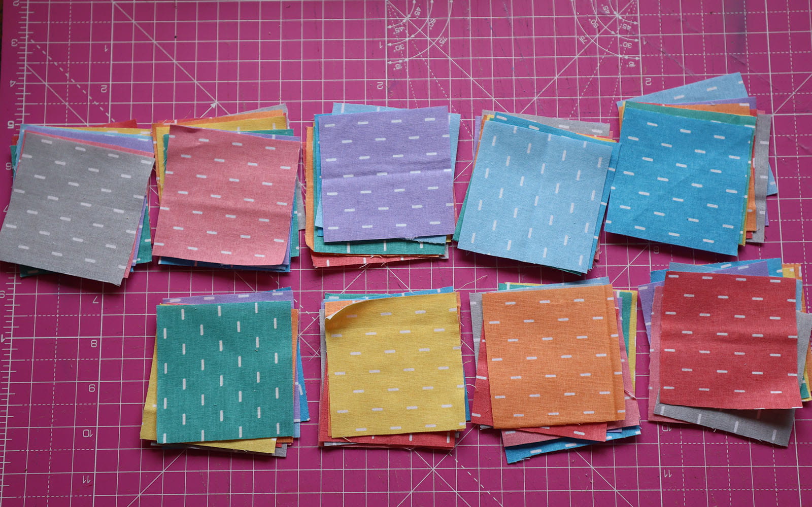 Nine stacks of sorted coloured fabric squares on pink cutting mat