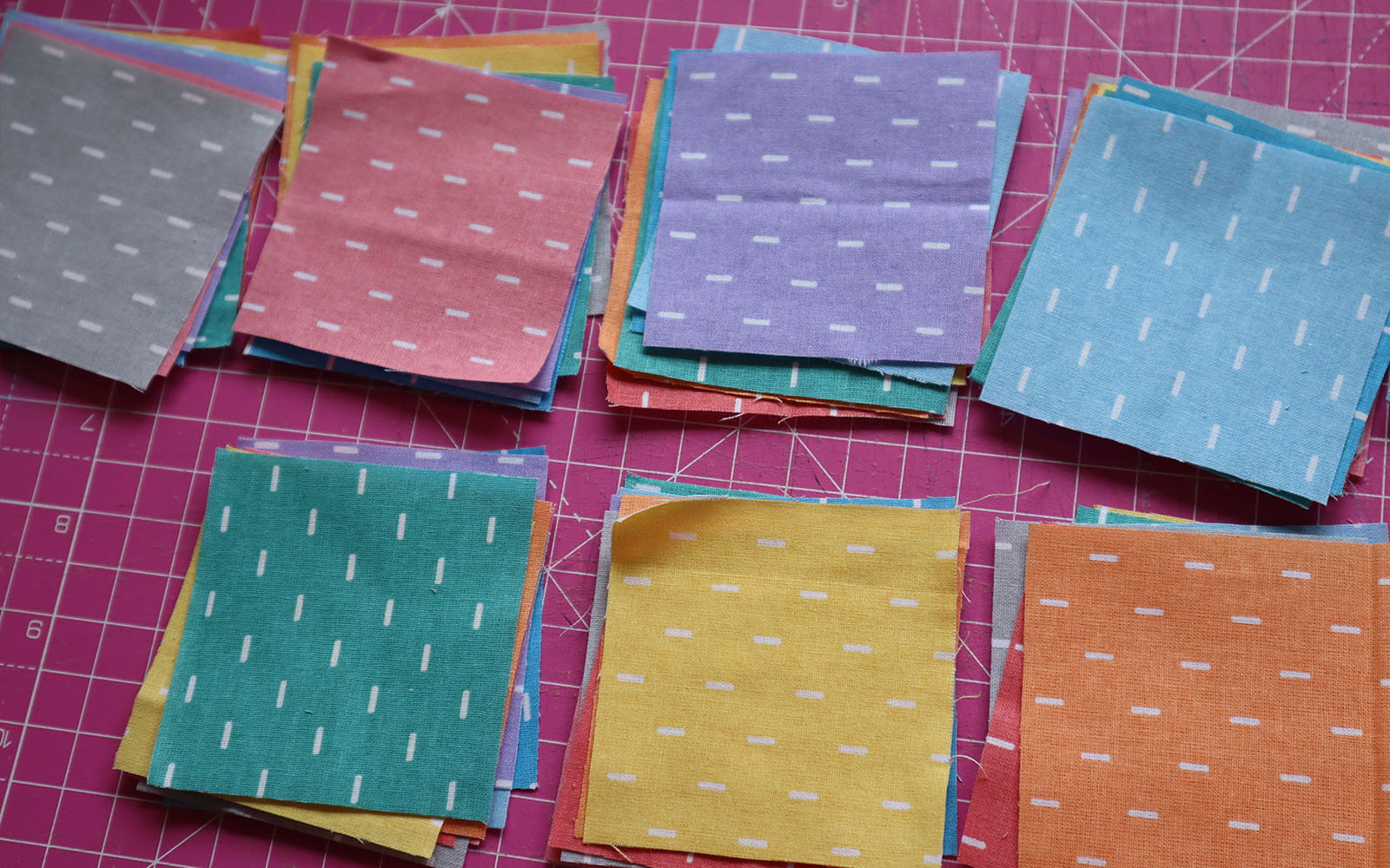 Seven stacks of sorted coloured fabric squares on pink cutting mat