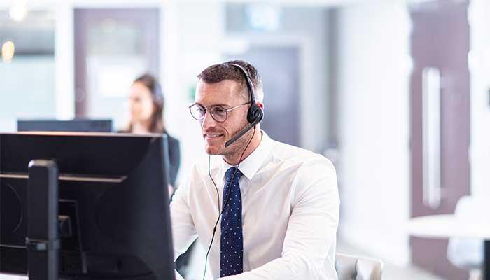 Male wearing shirt and tie in call centre environment sat by a computer with a headset on 